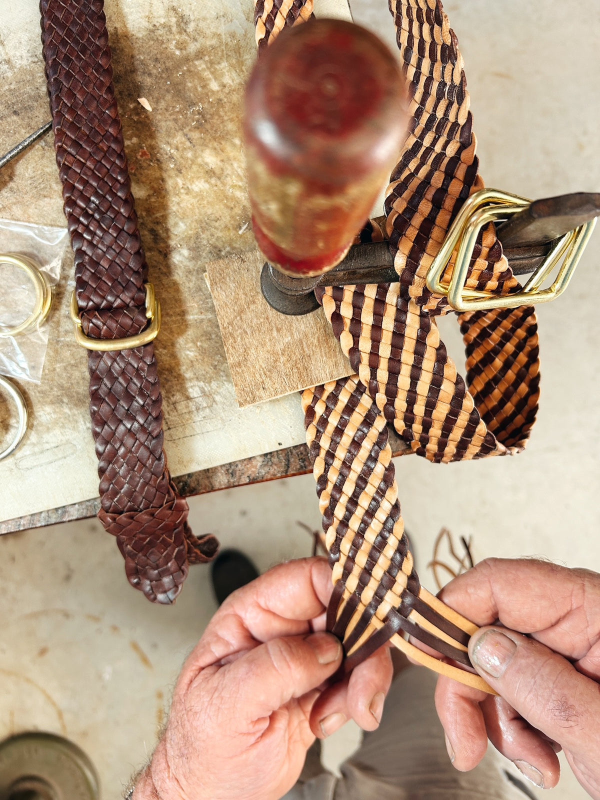 A close-up of a leather strap being woven or adjusted by hands in a workshop, with a matching brown leather strap and brass buckle hanging nearby on a workbench.